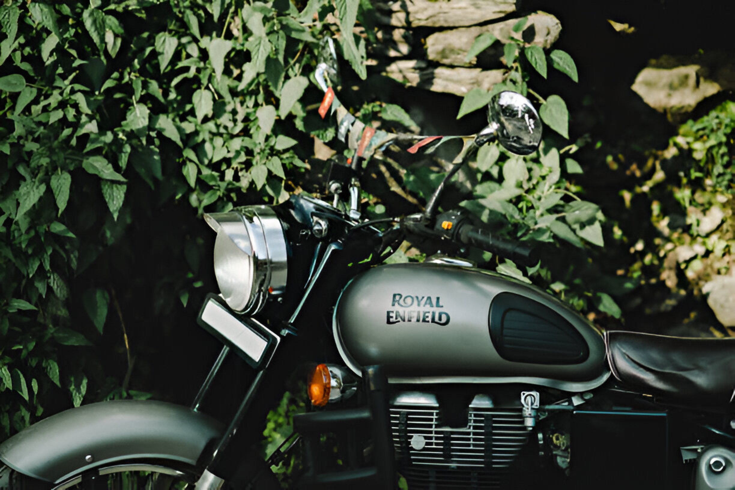 Classic Royal Enfield motorcycle with a 350-500cc engine, featuring a matte grey tank and chrome details, parked beside a green hedge.